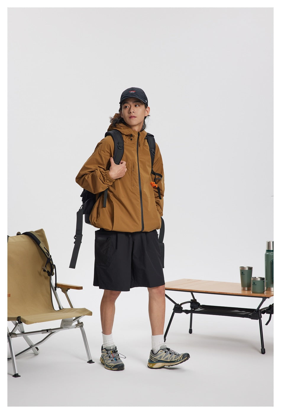 Model wearing a zipped mustard-brown hooded jacket with black shorts and a backpack.