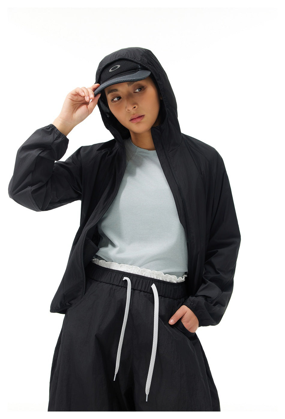 Model wearing a black hooded jacket over a pale blue shirt with loose black drawstring trousers and a cap.