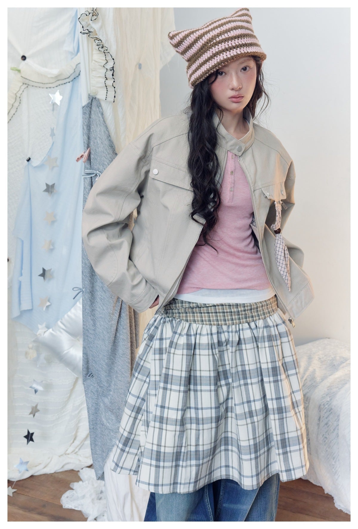 Model wearing an open light gray-beige utility jacket with flap pockets and a plaid hanging detail over a pink top.