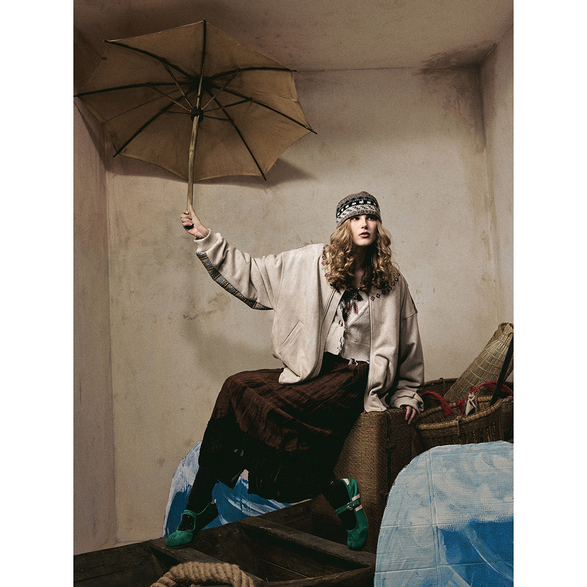 Model seated beneath an umbrella wearing an ivory jacket with patterned cuffs, a dark skirt, and a knit headband.