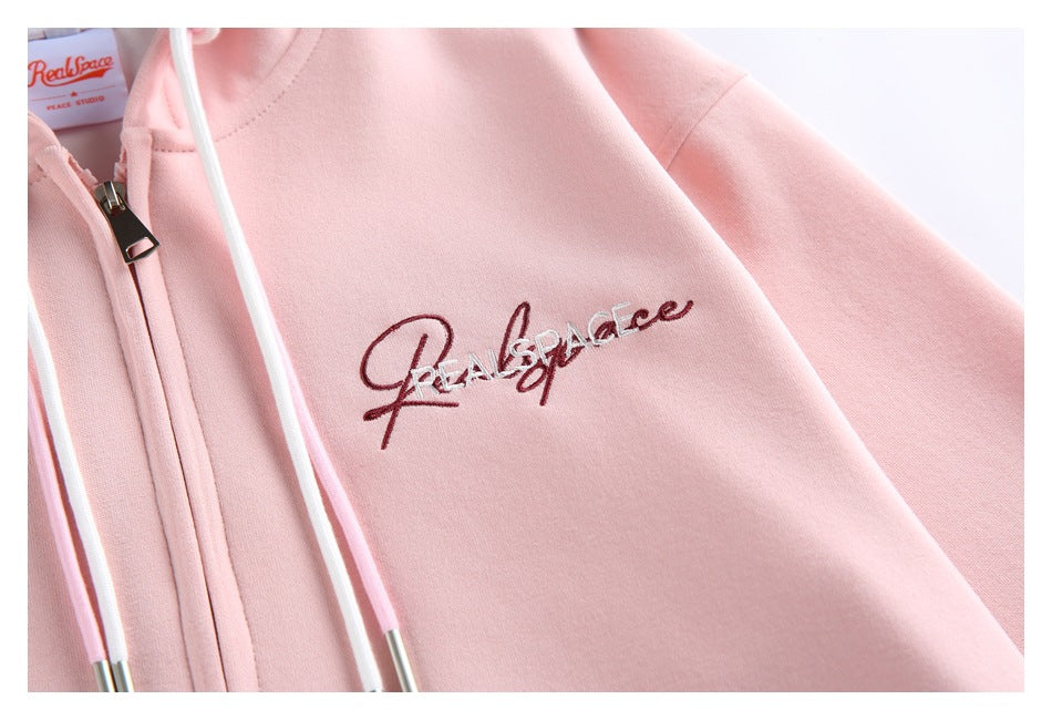 Close-up of dark script embroidery on the chest of a pale pink hoodie beside the zipper and drawstring.
