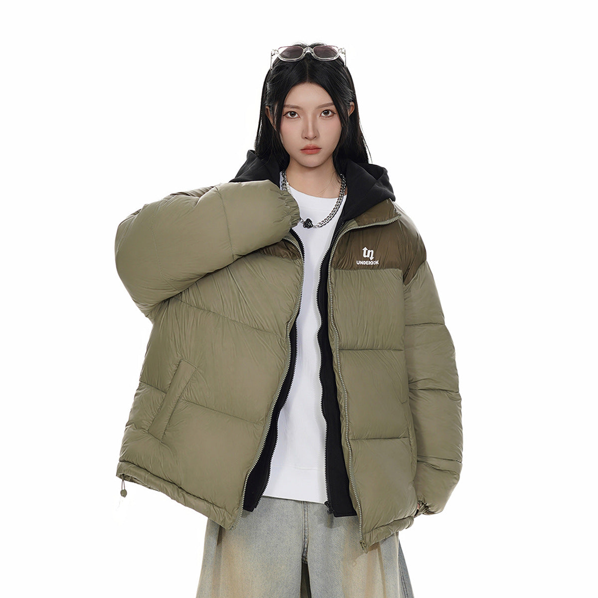 Model wearing an open olive puffer jacket with a dark brown shoulder panel, black hood, and chest logo.