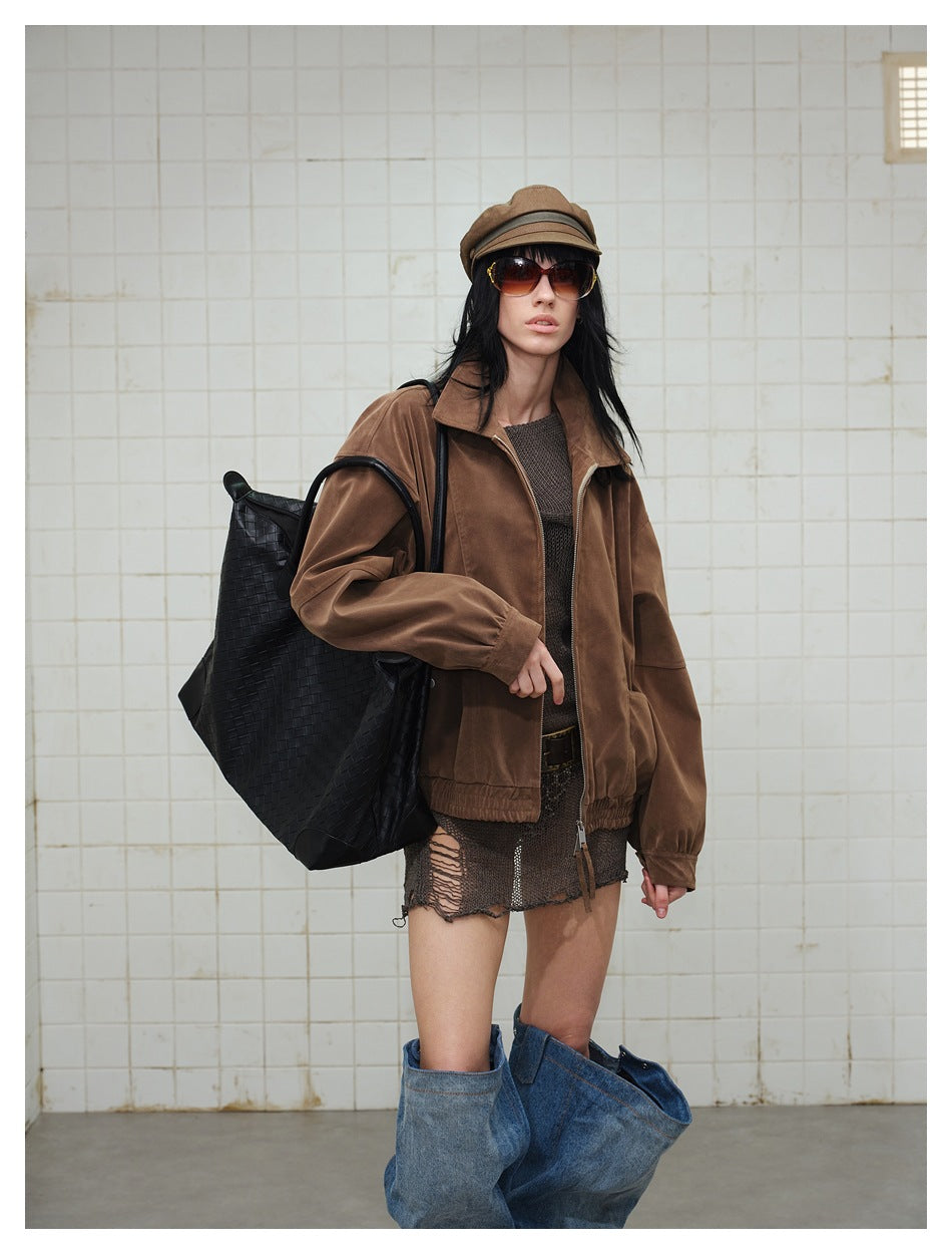 Model in a brown zip jacket carrying a large black woven tote, in a tiled room, three-quarter view