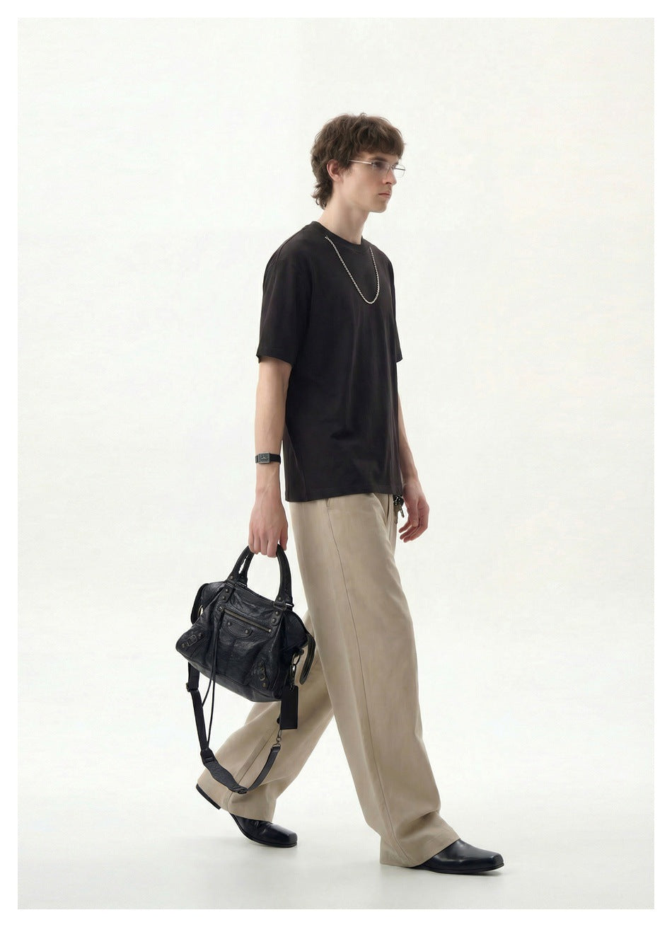 Man walking in a dark brown oversized T-shirt with a chain necklace, beige wide-leg pants and a black bag
