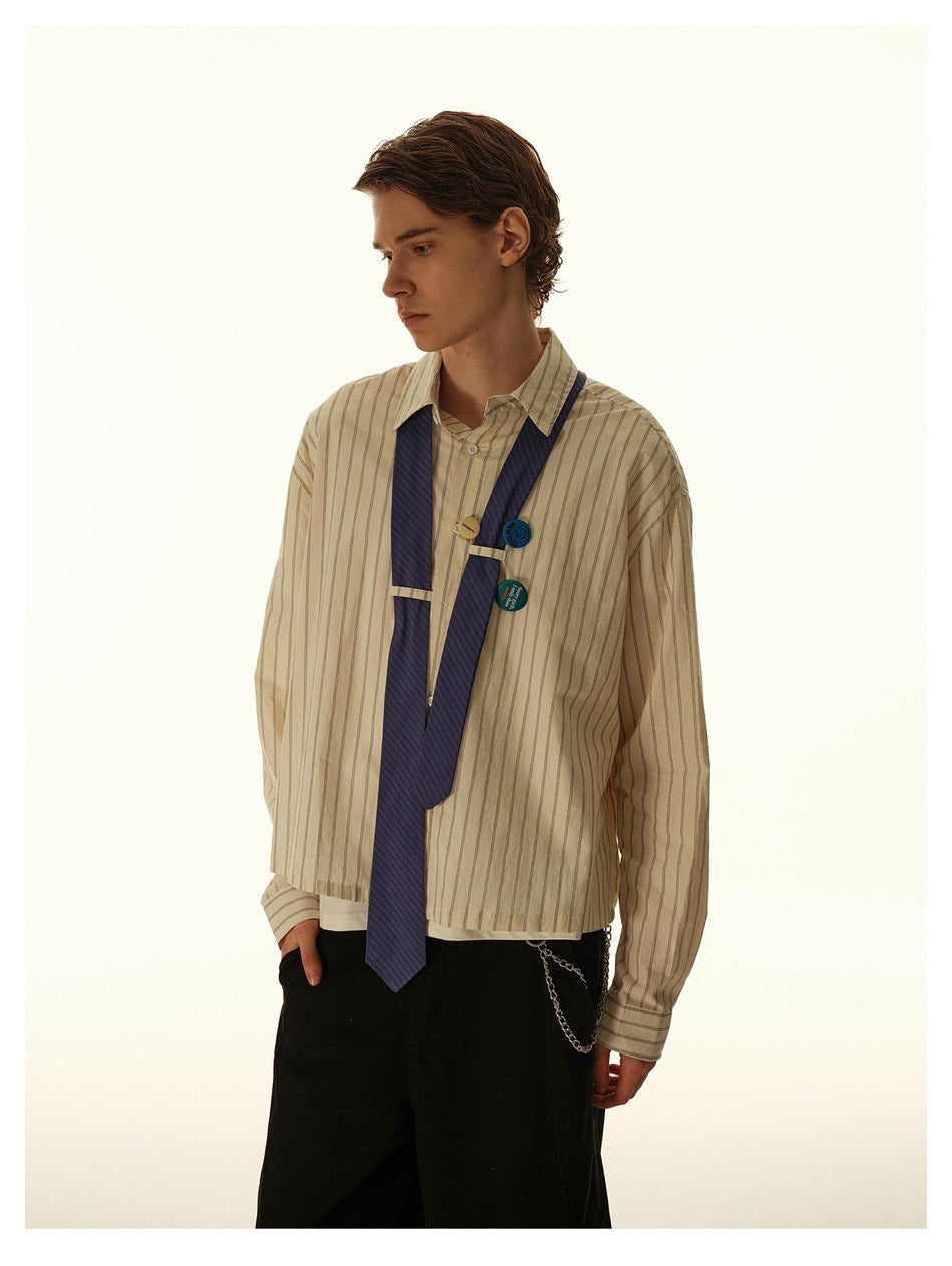 Male model in striped cream button-up shirt with blue tie draped over the chest, badge pins, black pants and chain