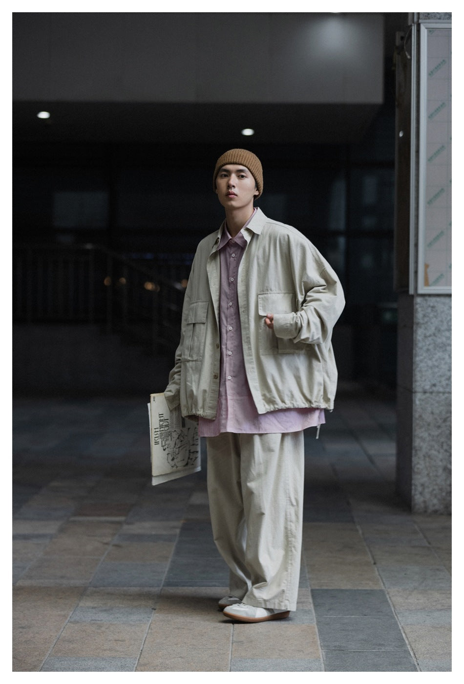 Model wearing a light beige oversized pocket jacket and loose trousers over a pink shirt, with a tan ribbed beanie.