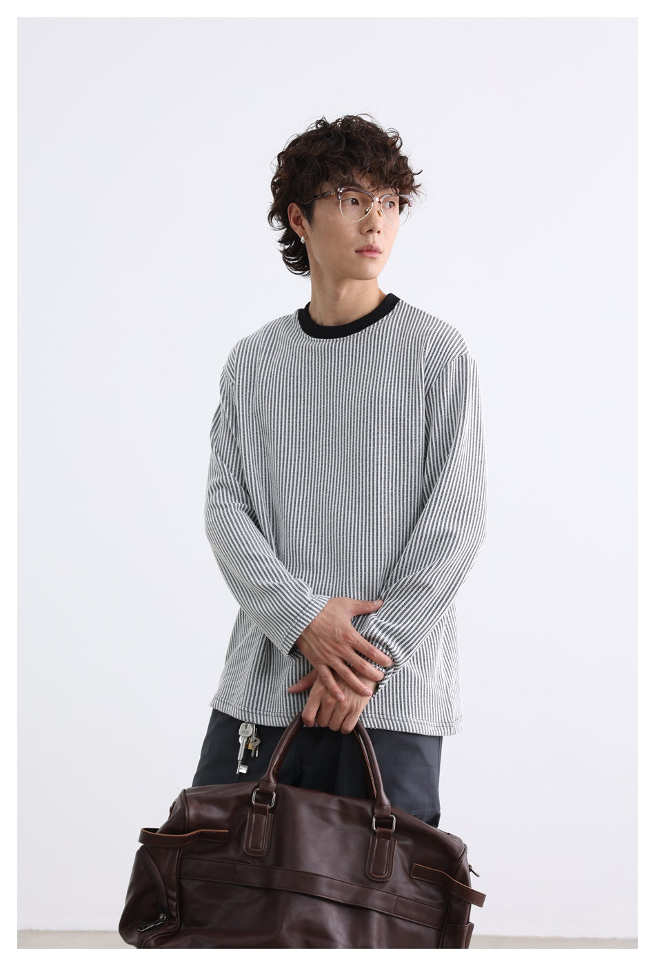 Model wearing a gray-and-white vertically striped long-sleeve shirt with a black crew neck, holding a brown bag.