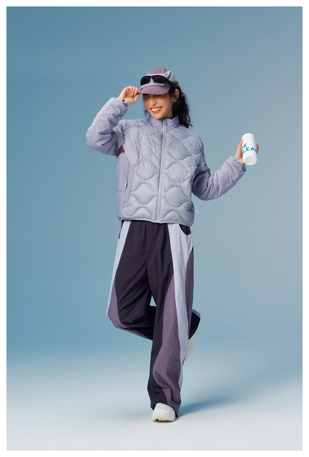 Woman modeling a pale lavender wave-quilted zip jacket with dark color-block pants and a cap.