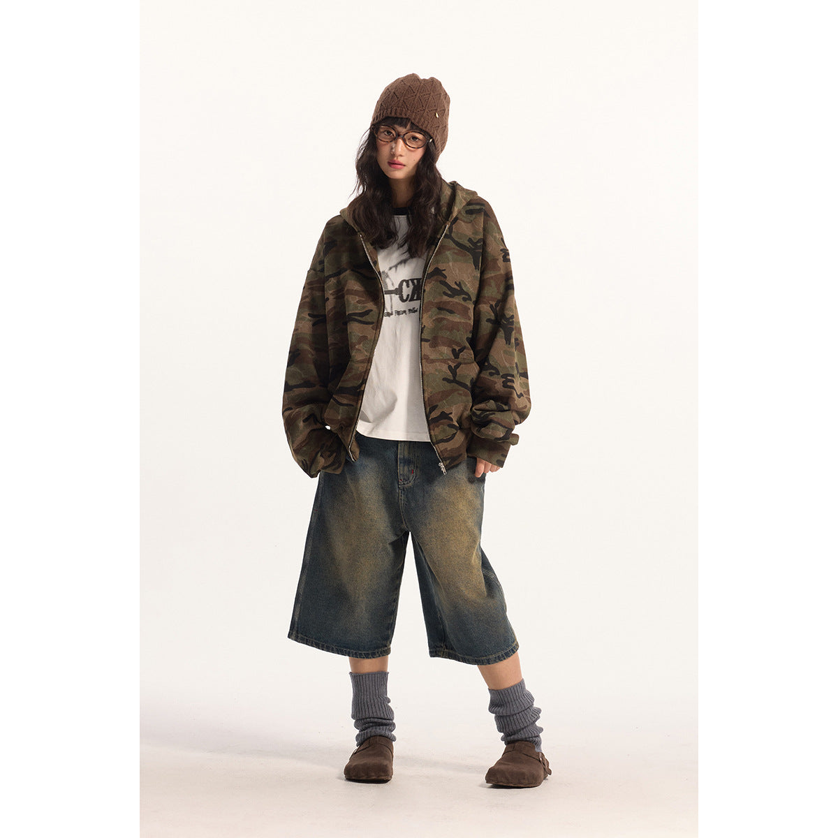 Model wears an open camouflage zip jacket over a white graphic tee and wide dark denim shorts.