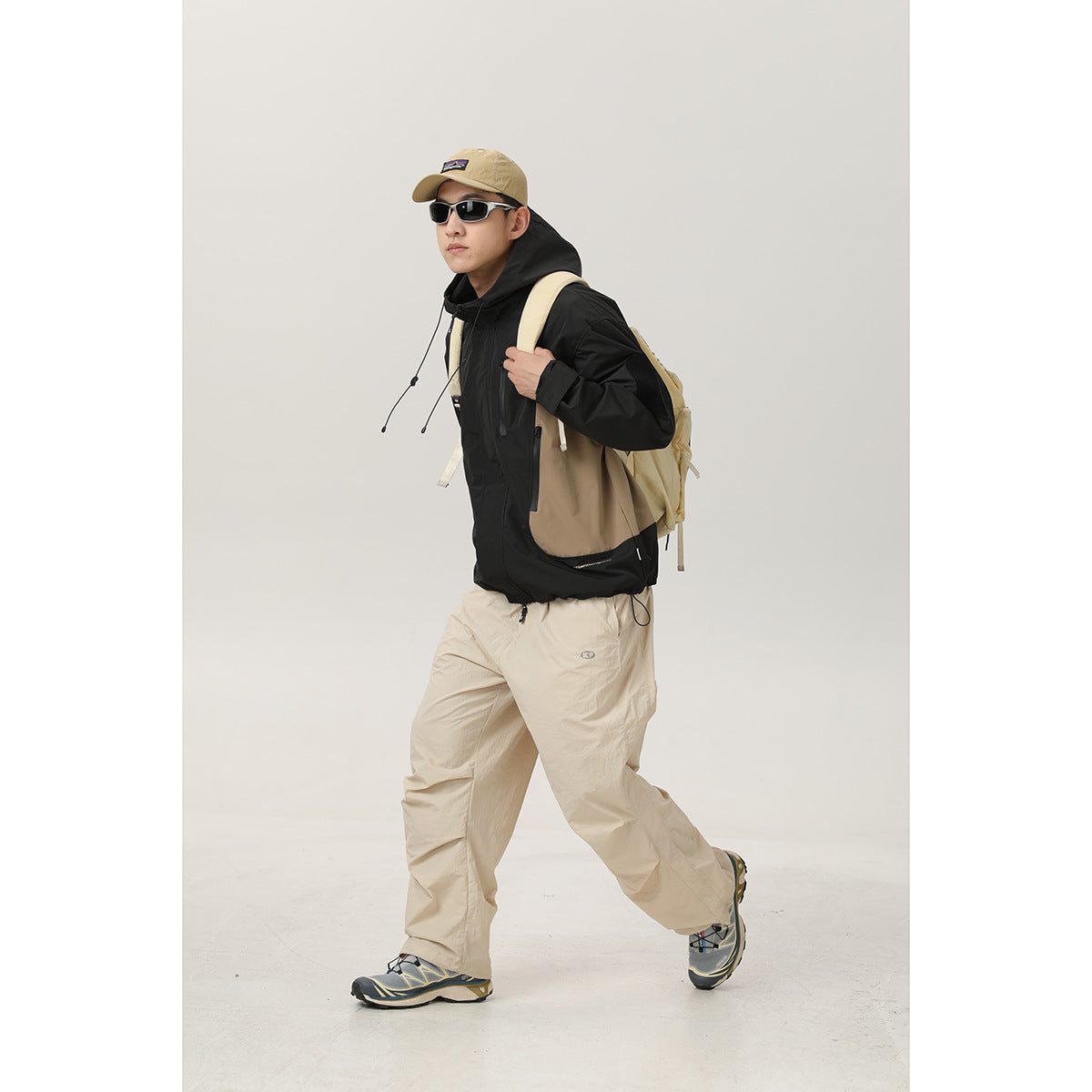Full-length model walking in a black and tan hooded jacket, beige trousers, cap, and backpack.