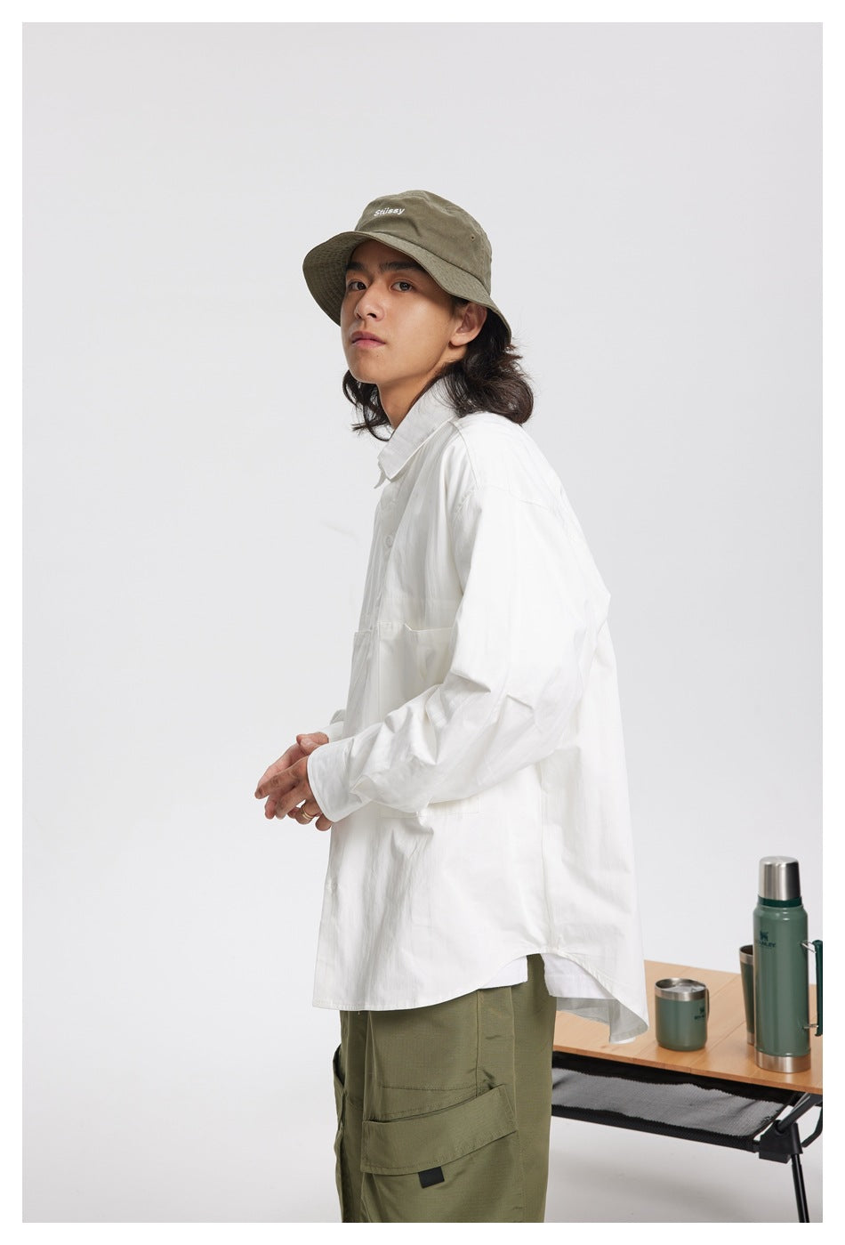 Side view of a model wearing a loose white long-sleeve button-up shirt with a curved hem and olive cargo pants.