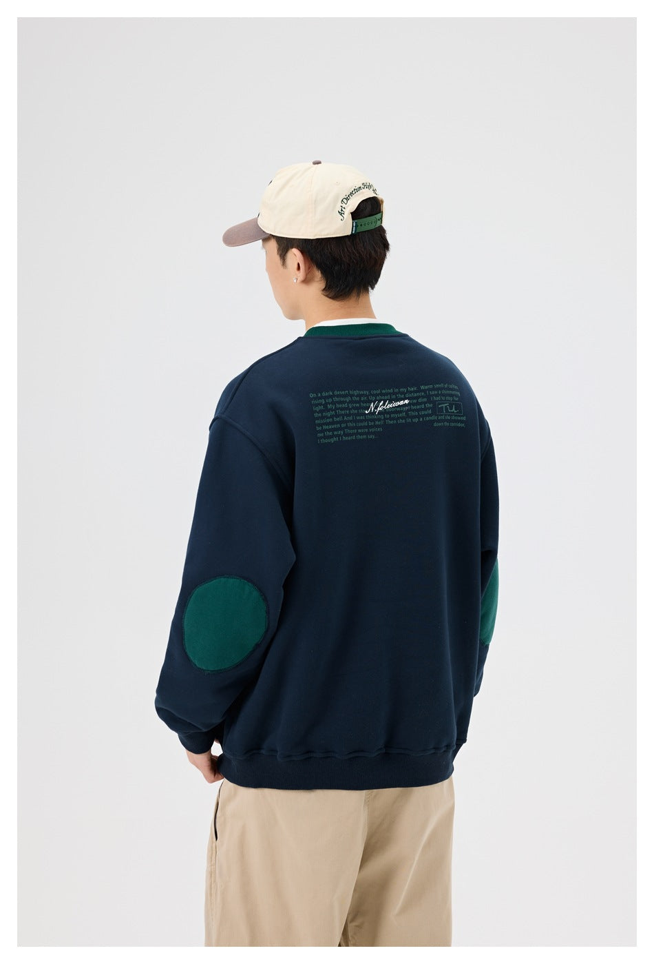 Back view of a model wearing a navy sweatshirt with green elbow patches and small text across the upper back.
