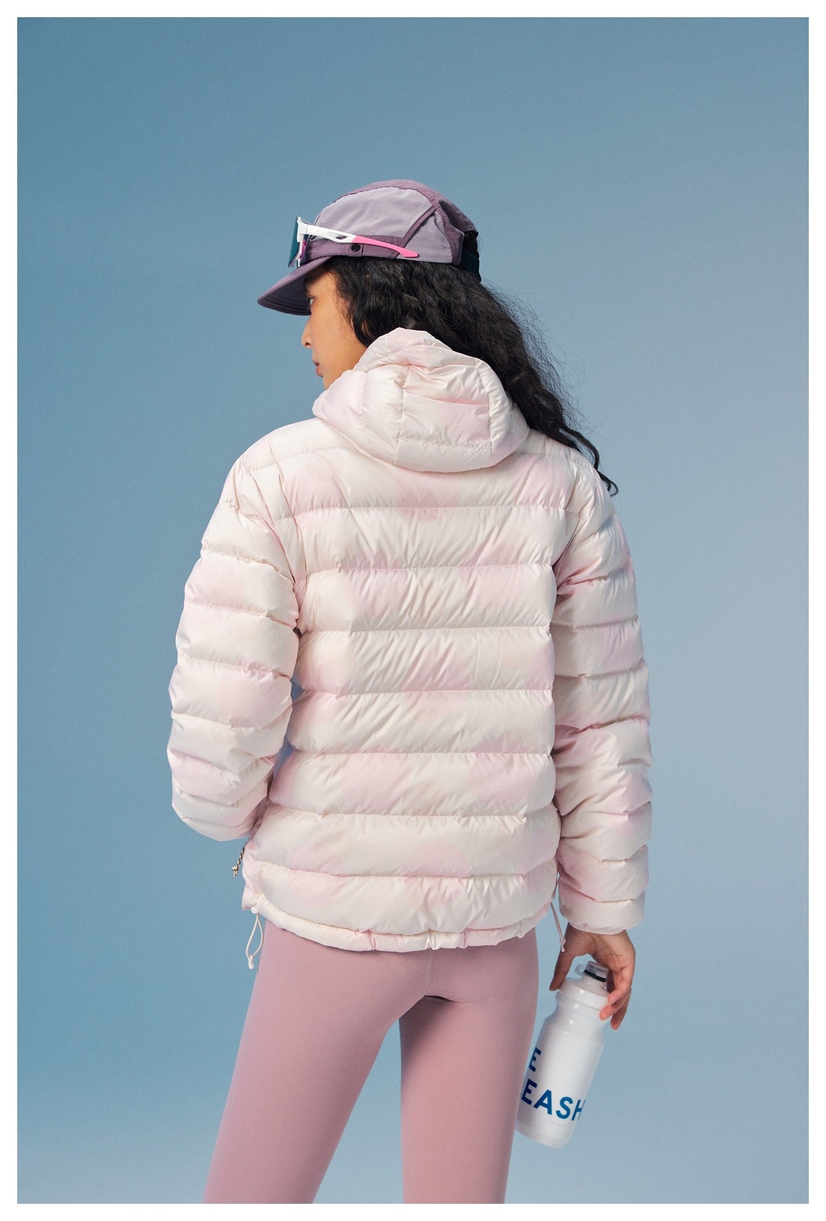 Rear view of a model wearing a pale pink hooded puffer jacket and lavender leggings, holding a water bottle.