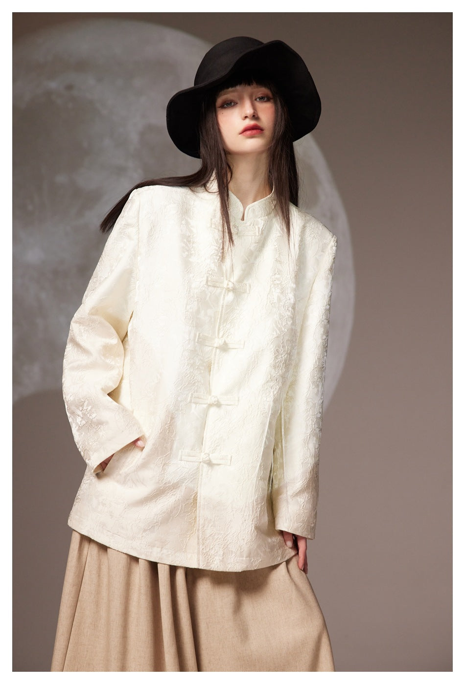 Front view of a model in an ivory floral-textured jacket with frog closures, black hat, and beige skirt.