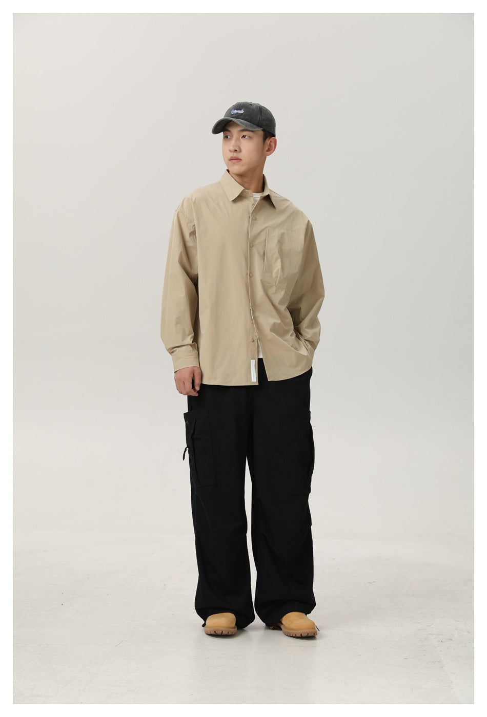 Model wearing a khaki button-up shirt open over a white tee with black cargo pants, a gray cap and tan boots