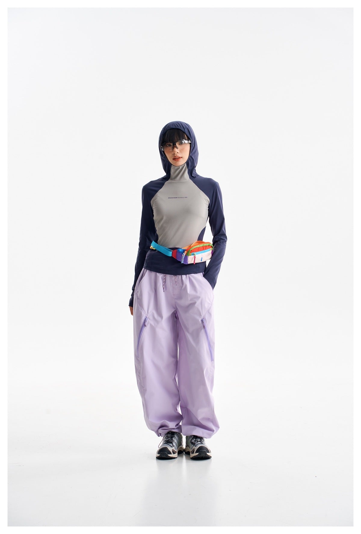 Full-length model wearing a navy and gray hooded top, loose lavender pants, and a colorful waist bag.