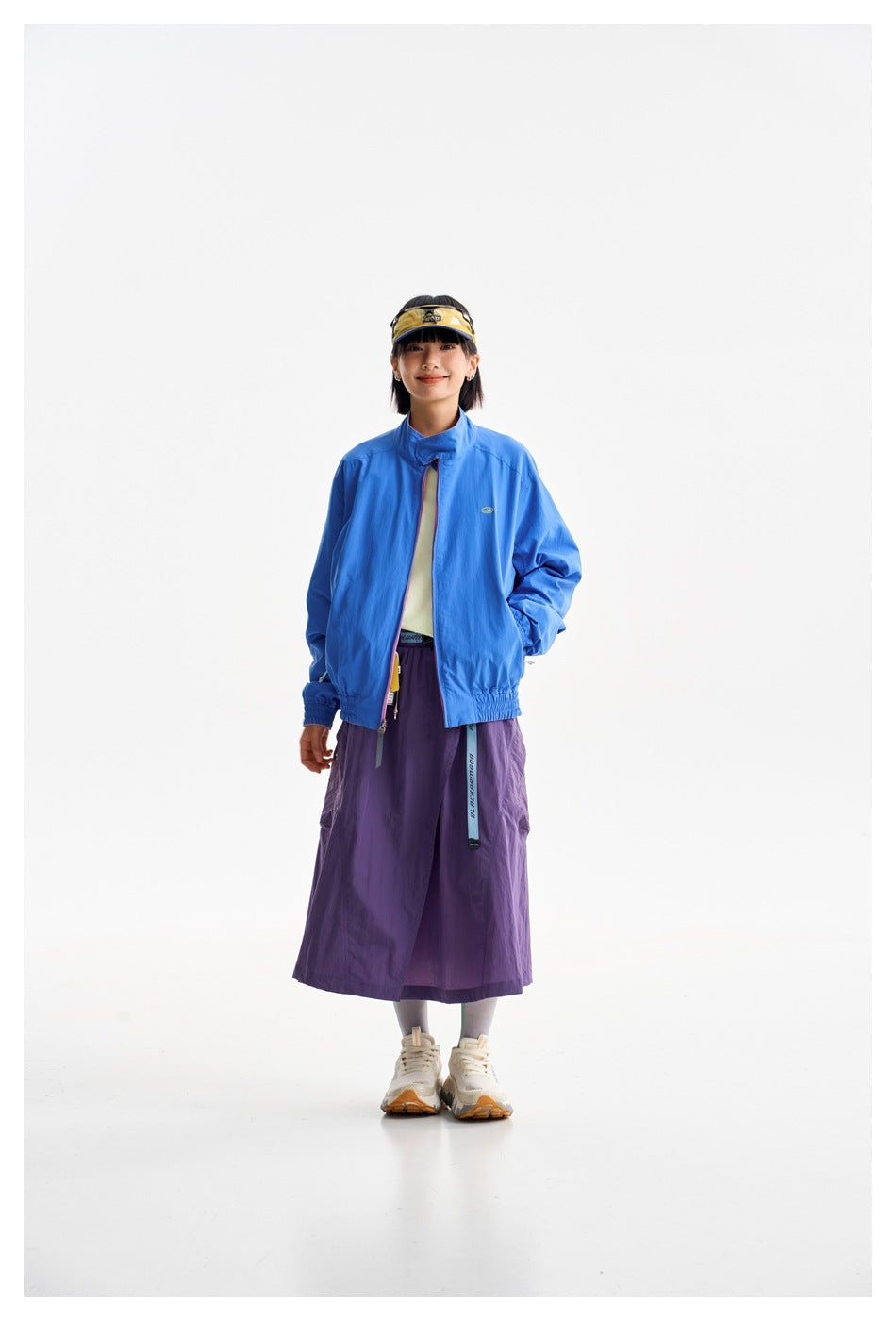 Full-length model wearing a bright blue zip jacket and long purple skirt with a yellow visor and sneakers.