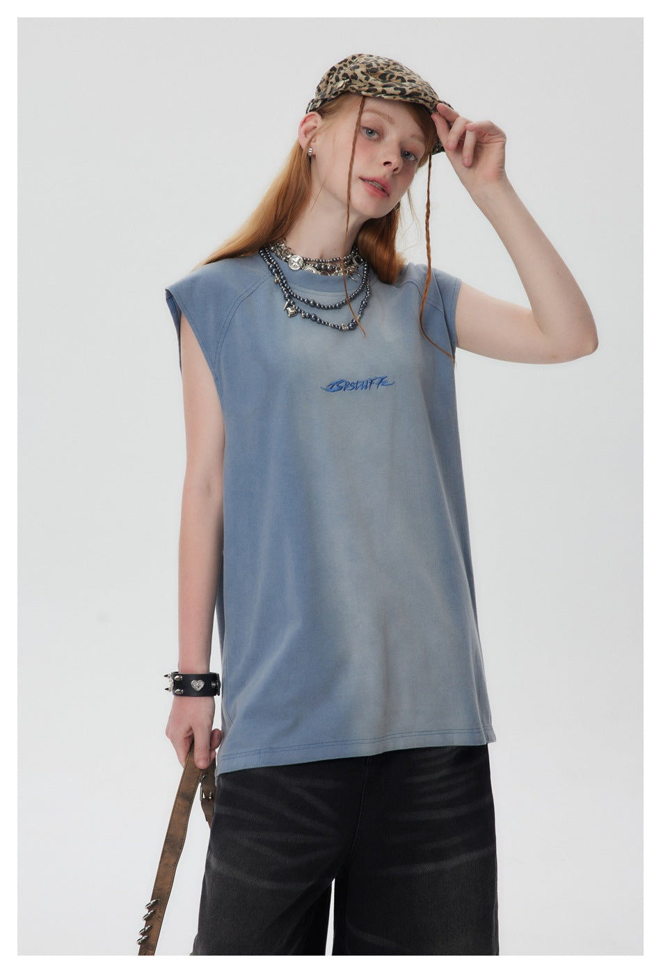 Model wearing a faded blue sleeveless crew-neck top with chest embroidery, layered necklaces and a leopard cap
