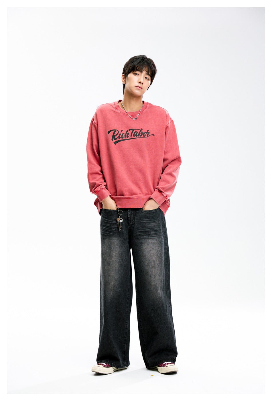 Model wearing relaxed red washed sweatshirt with black script chest print, hands in jeans pockets, front view
