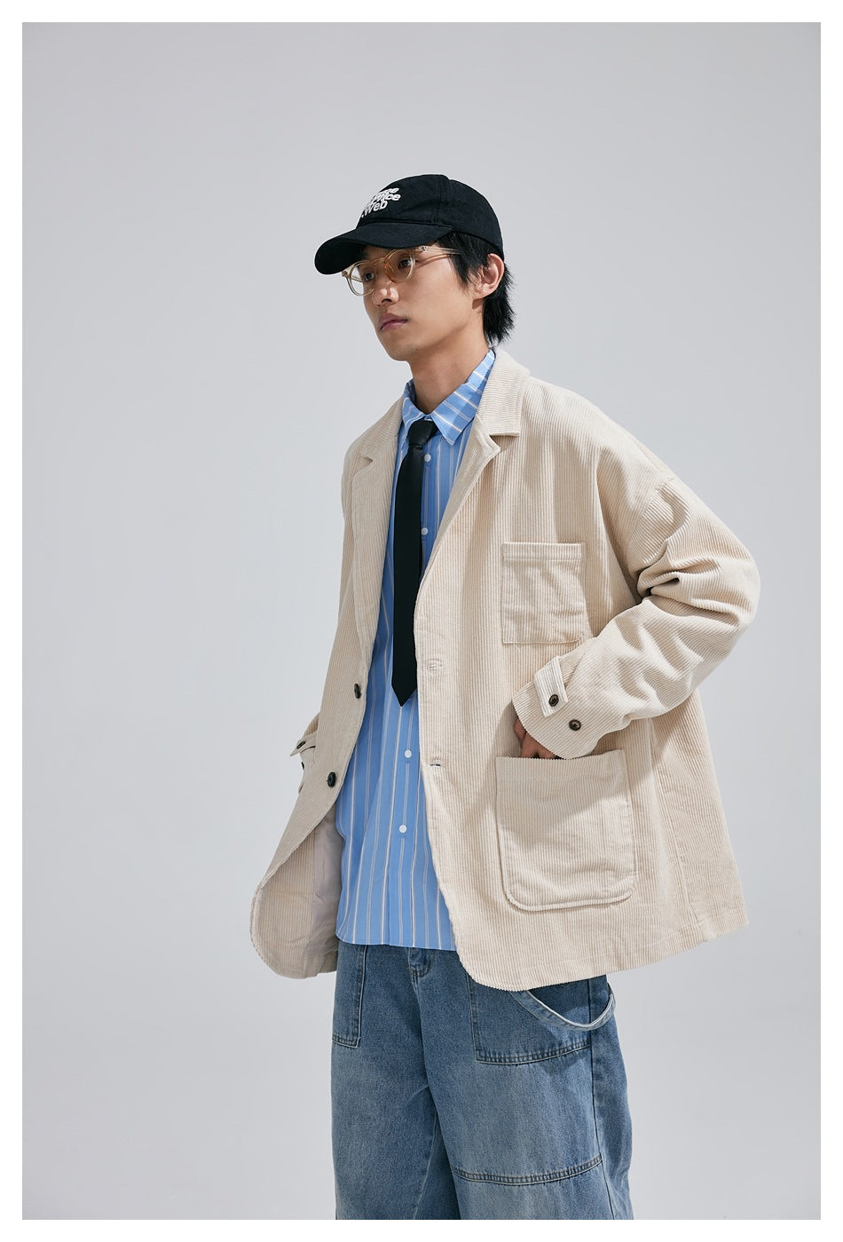 Model wearing an oversized cream corduroy blazer with a blue striped shirt, black tie, and loose jeans.