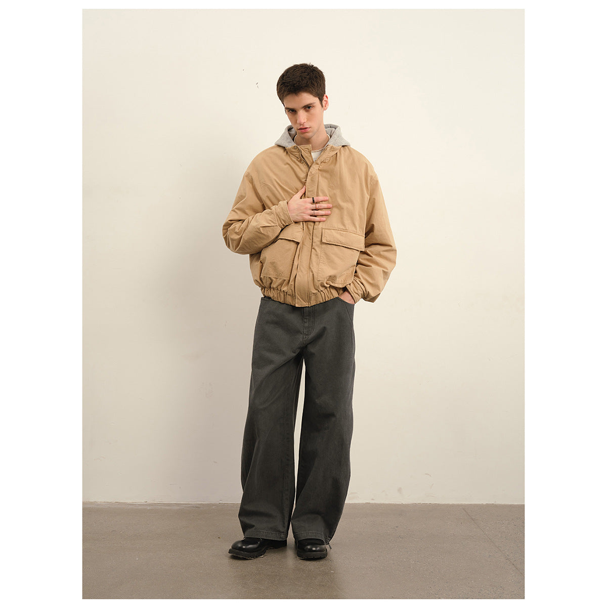 Model wearing a tan hooded jacket with large flap pockets and wide charcoal trousers.