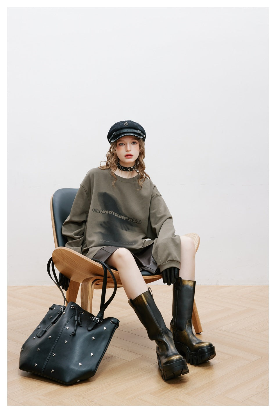 Model seated in an olive-gray oversized graphic sweatshirt, shown with a black studded tote and tall boots.