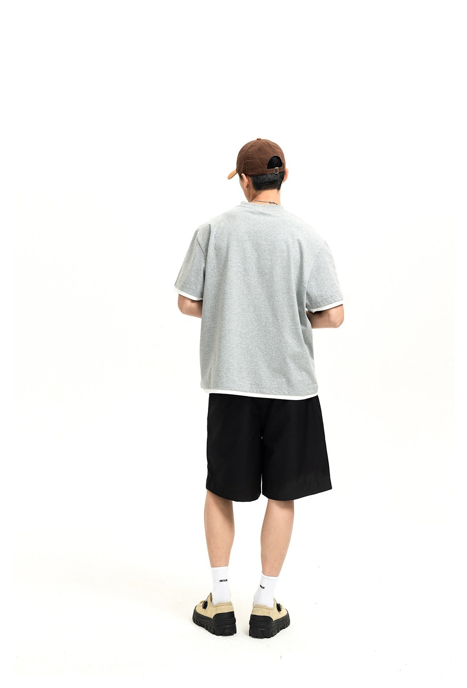 Back view of a man in a heather gray T-shirt with white layered hem, black shorts and a brown cap