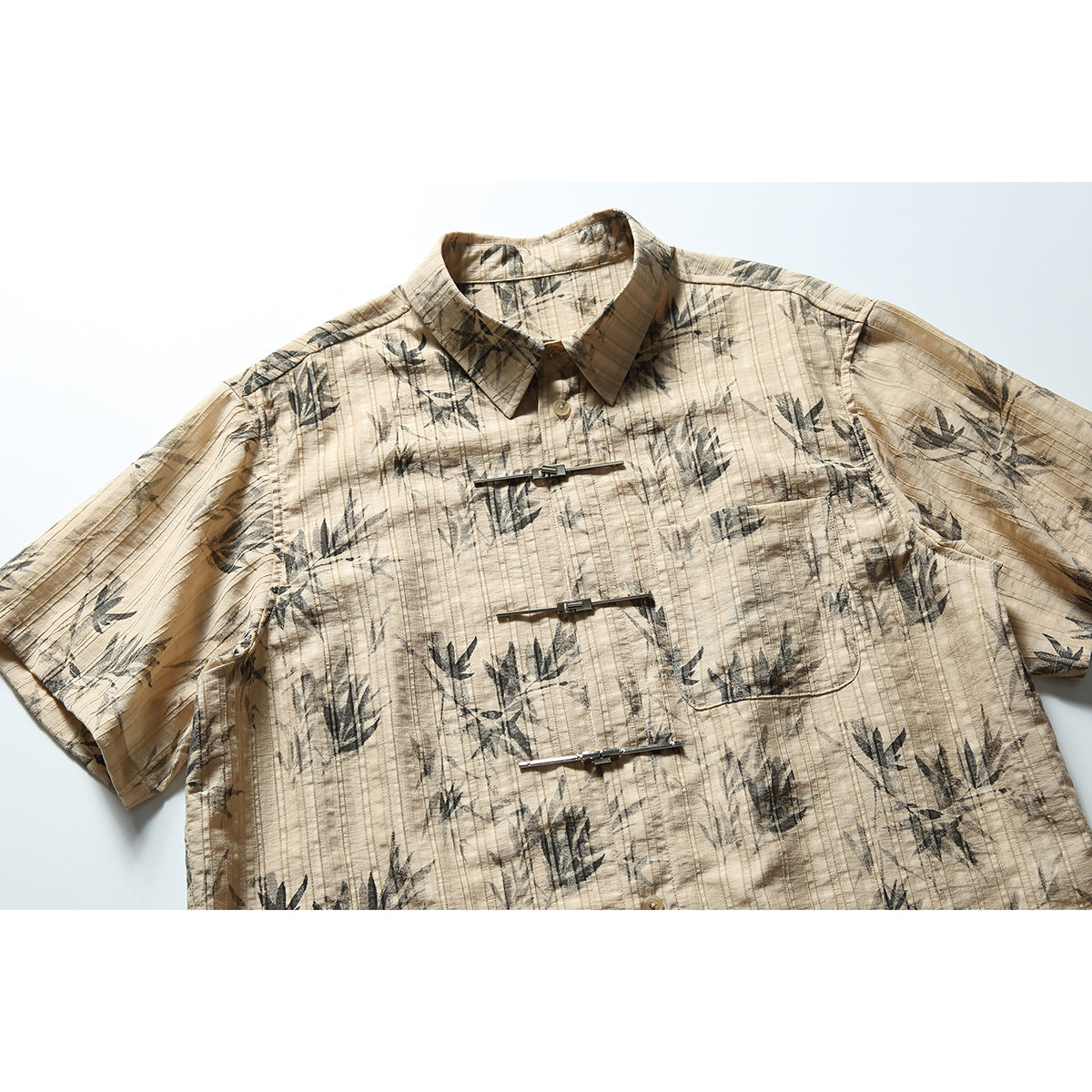 Front view of a tan short-sleeve collared shirt with a dark botanical print, chest pocket, and three silver clasps.