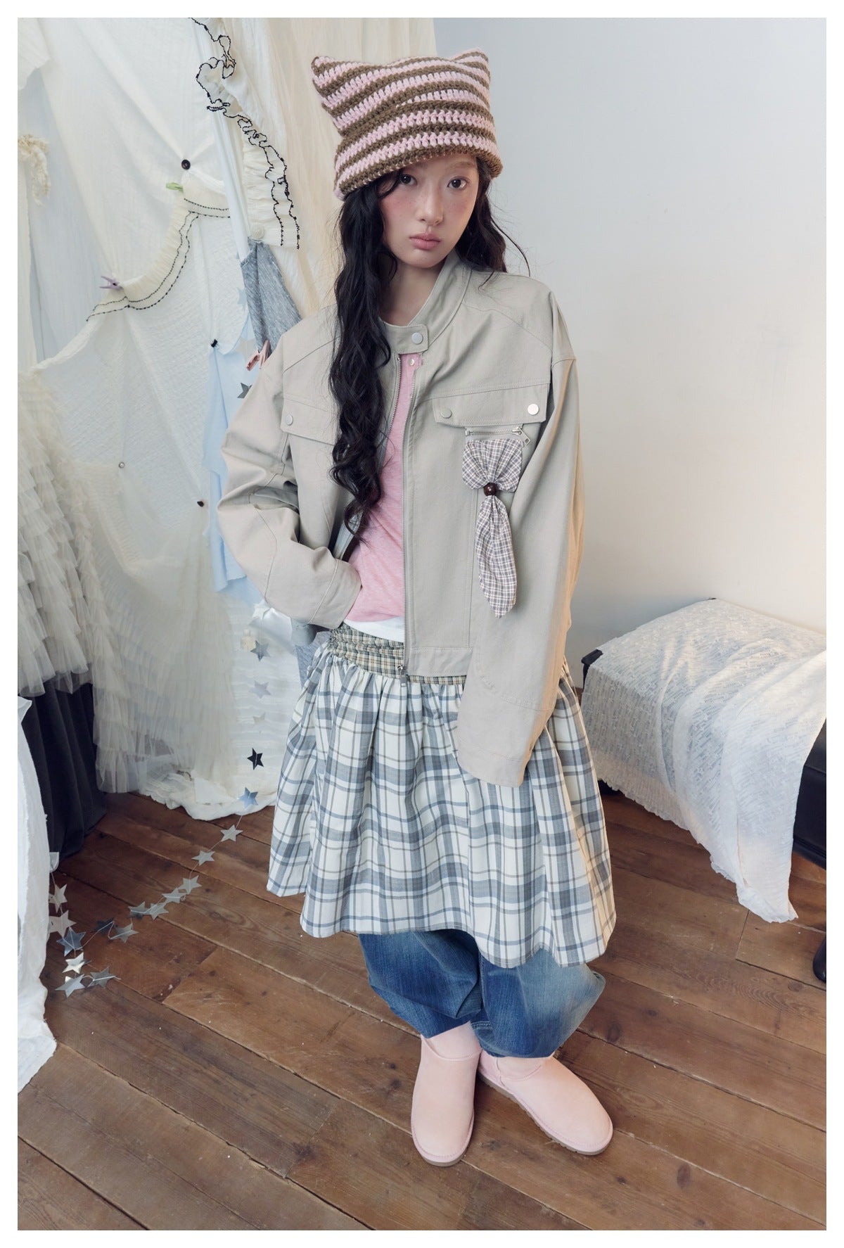 Model wearing a light gray-beige utility jacket with flap pockets and a plaid hanging detail, layered over a pink top.