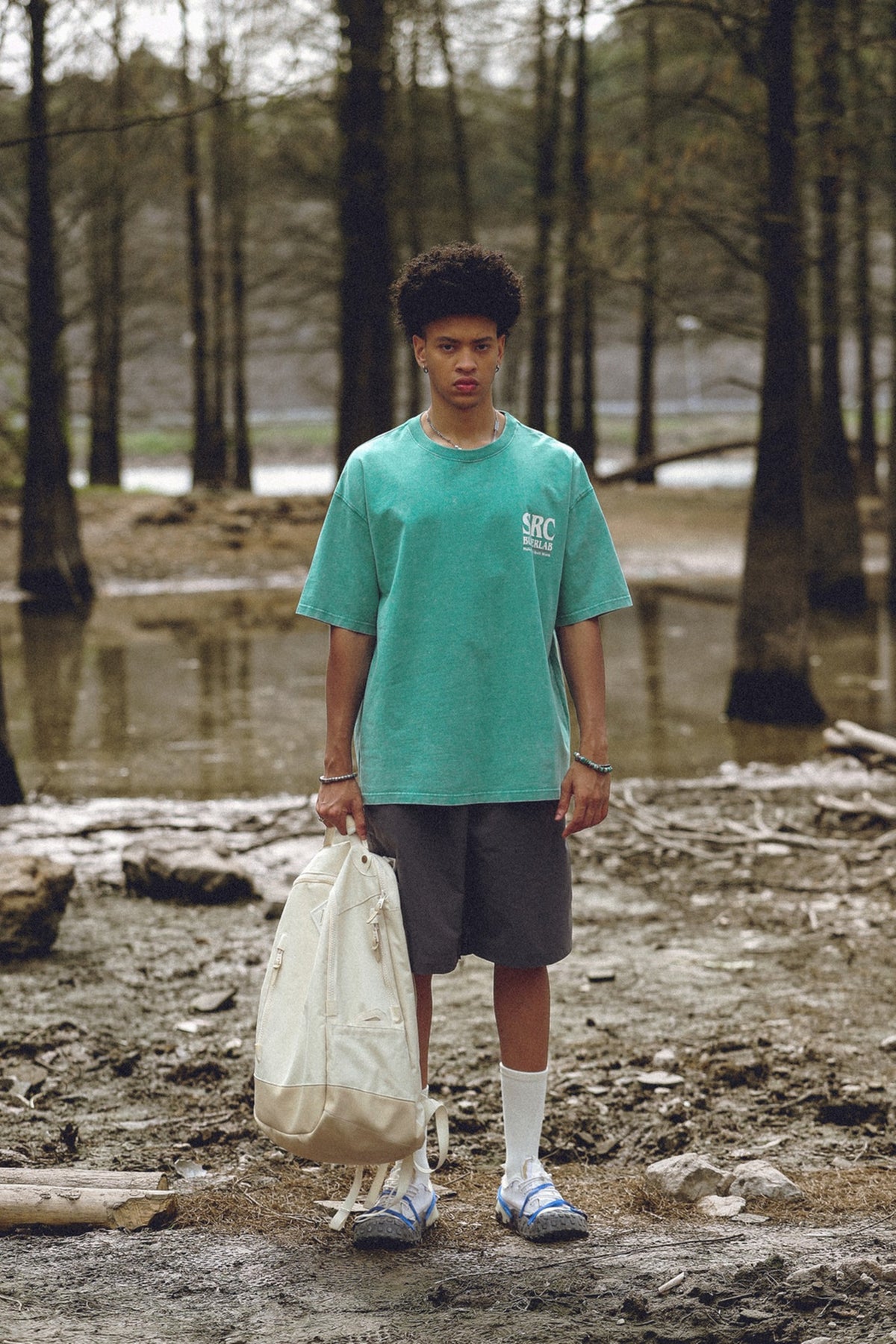 Model wearing a teal acid-wash graphic T-shirt with dark shorts, standing outdoors.