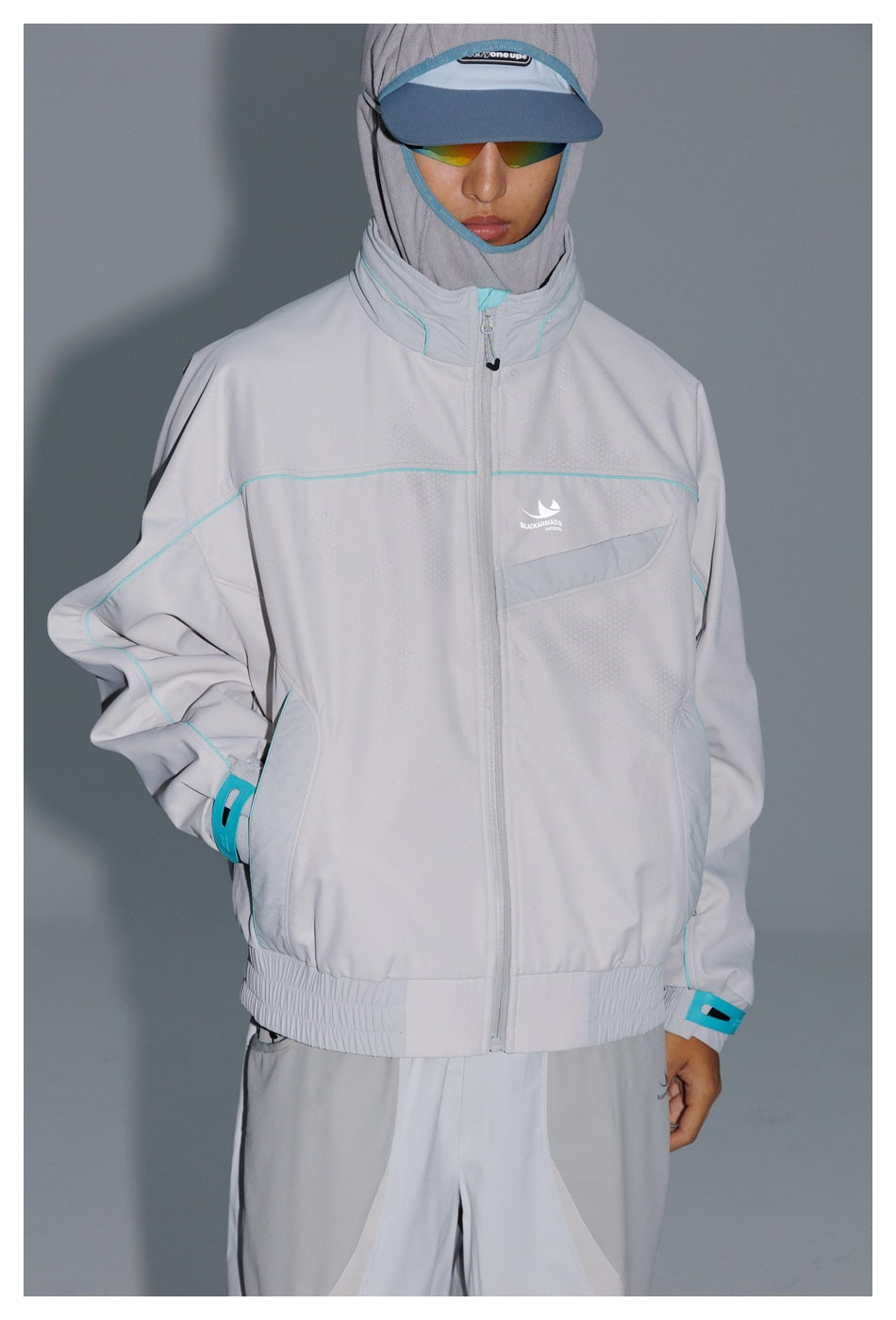 Model wearing a light gray zip-up jacket with turquoise piping and matching pants, cap, and reflective sunglasses.