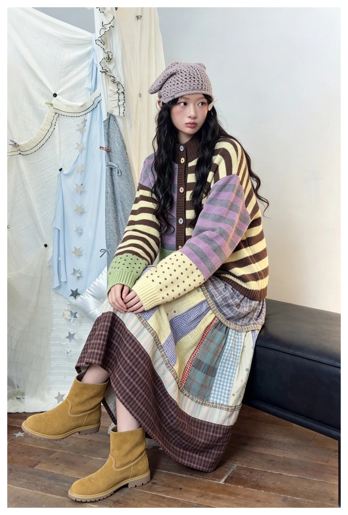 Woman seated on a bench wearing a striped colorblock cardigan with a patchwork skirt, boots and a knit headscarf