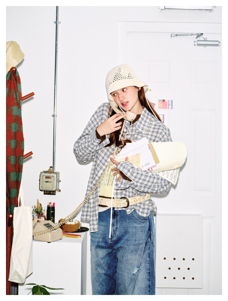 Model wearing a blue-gray plaid button-up shirt and cream crochet hat while holding a telephone and papers.