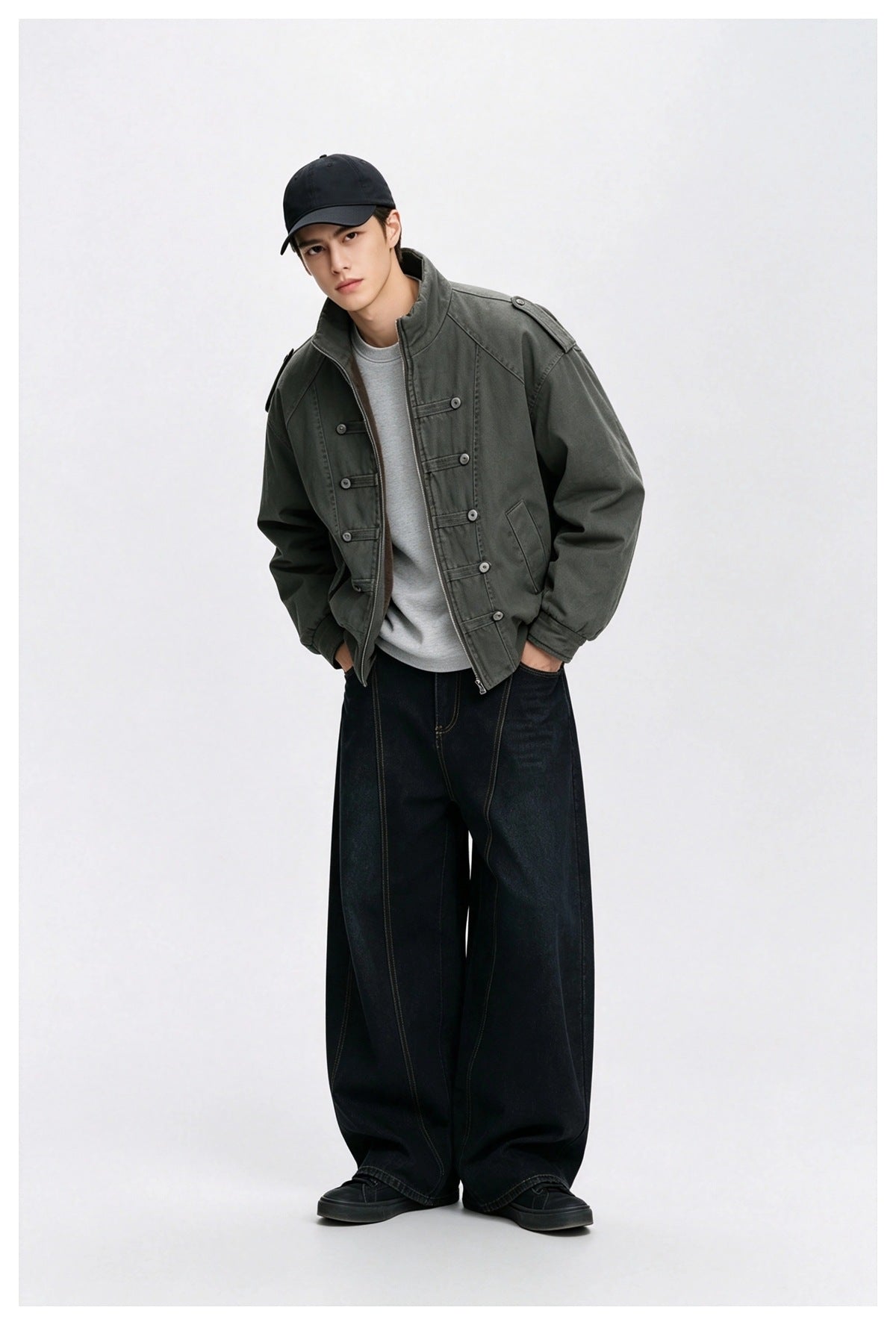 Man wearing the olive green stand-collar jacket open over a gray sweatshirt with black wide-leg jeans and a dark cap