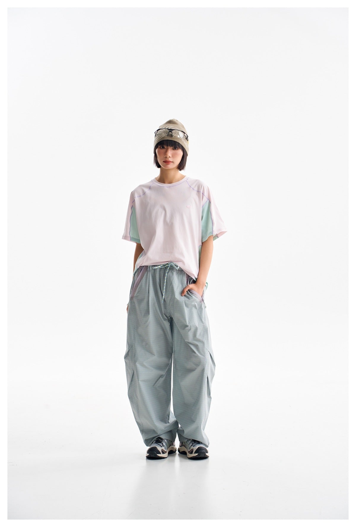 Model wearing loose sage-check trousers and a white pastel-panel T-shirt, with a beanie and sneakers.