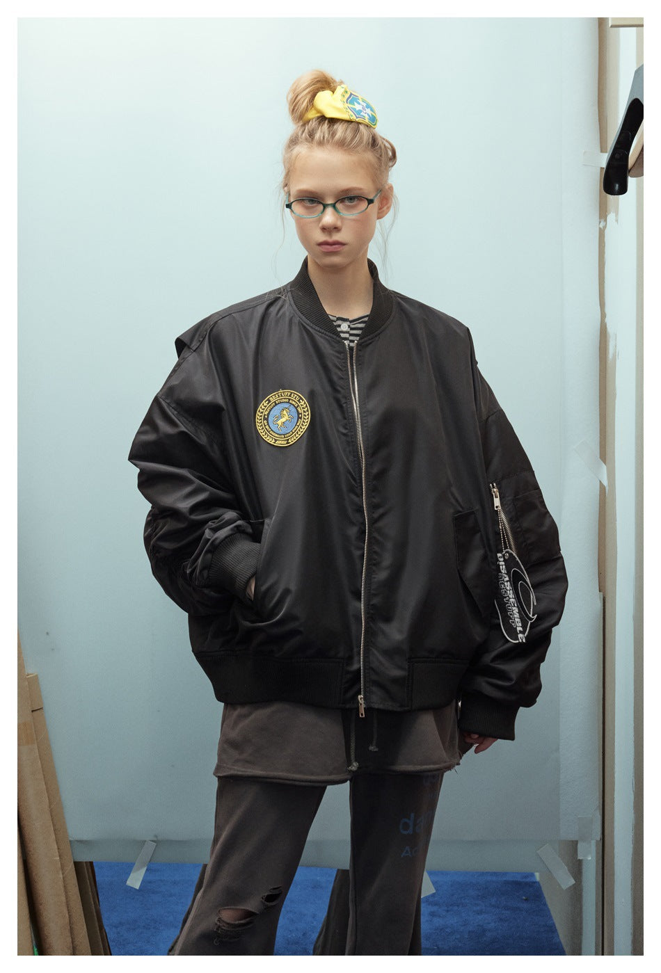 Closer front view of a model wearing an oversized black bomber jacket with a chest patch and zippered sleeve pocket.