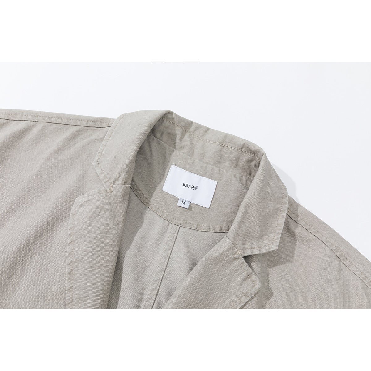 Close-up of a light gray-beige jacket's notched lapel and sewn neck label marked BSAPA, size M.