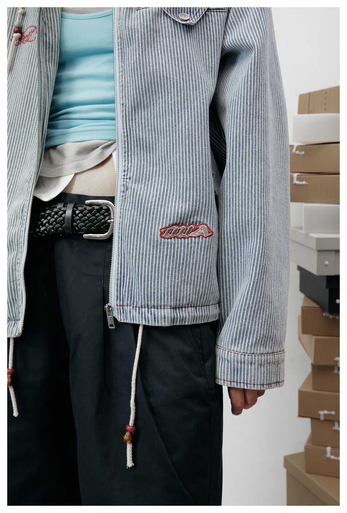 Cropped view of a model wearing a blue-and-white pinstripe zip jacket with red stitching and embroidered patches.