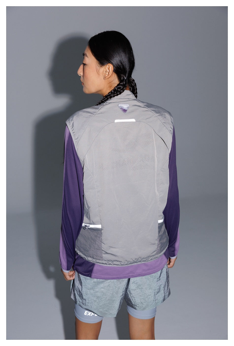Rear view of a model wearing a light gray sleeveless vest over a purple top, with gray shorts.