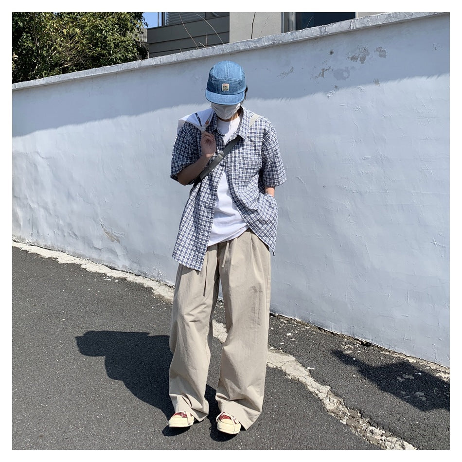 Person wearing khaki wide-leg trousers with a plaid short-sleeve shirt and denim cap, standing by a white wall