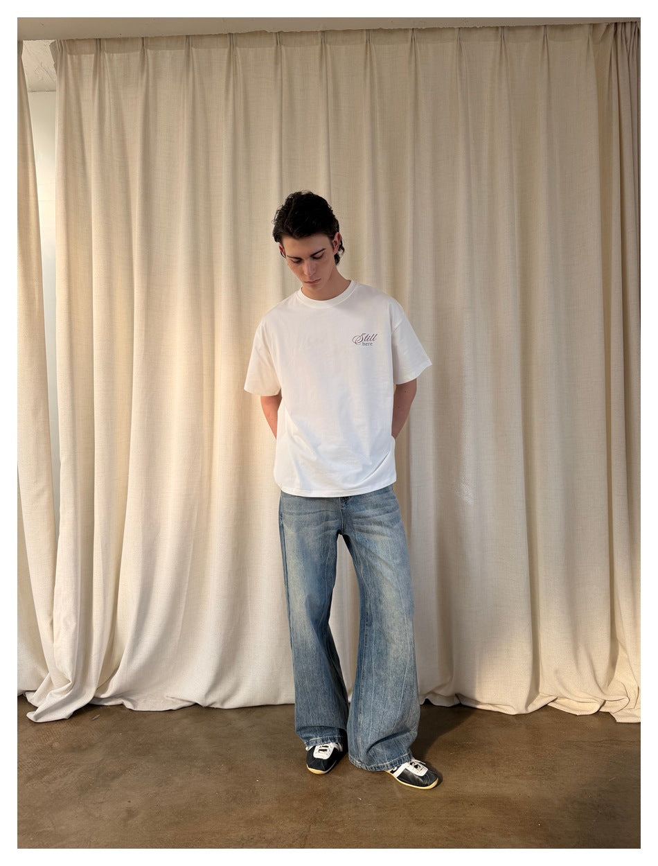 Model in blue faded wide-leg jeans, white T-shirt and black sneakers standing in front of a cream curtain