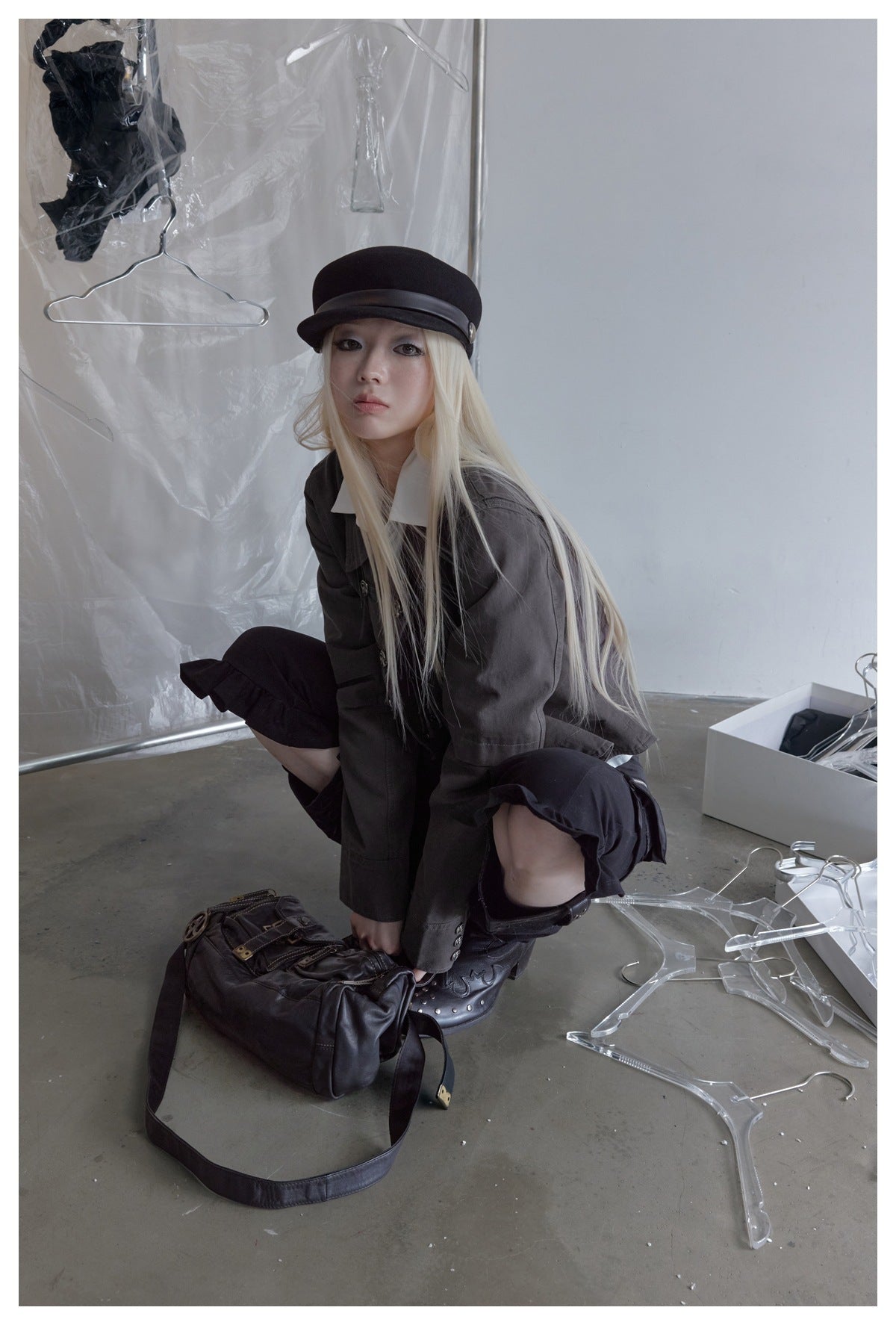 Crouching model in a charcoal jacket and black ruffled trousers, with a black cap and boots.