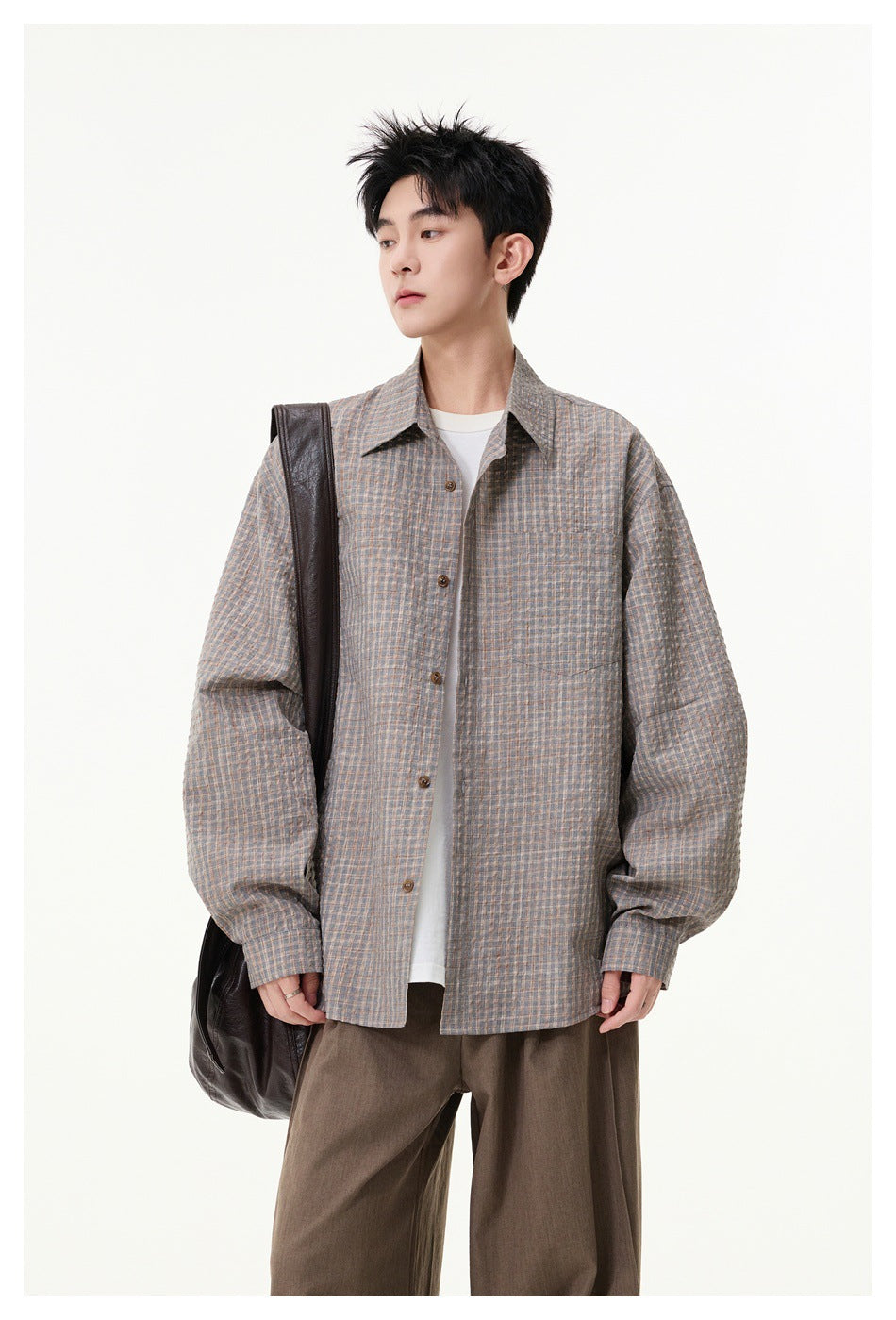 Model wearing a gray plaid textured button-up shirt open over a white tee with brown trousers and a dark shoulder bag