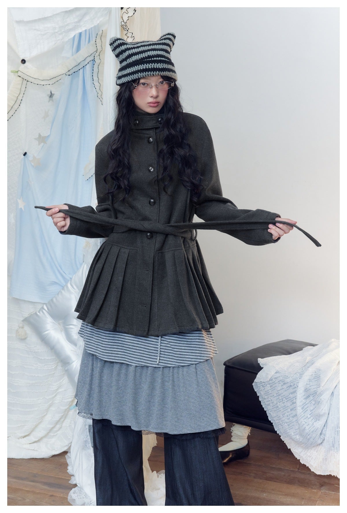 Model displays a charcoal button-front coat with a high collar, flared pleated hem, and long belt over a striped skirt.