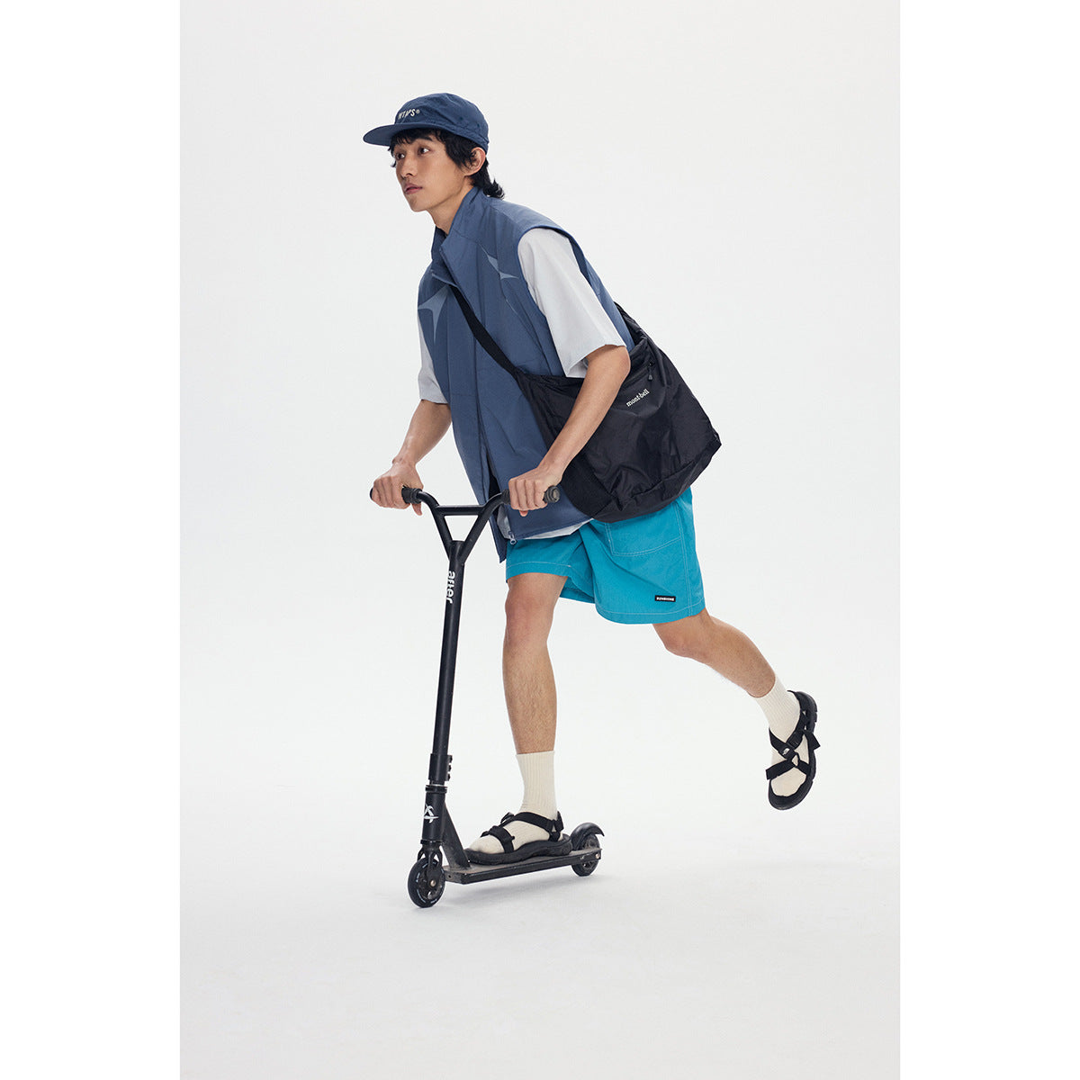 Model riding a scooter in a slate-blue vest, white T-shirt, turquoise shorts, navy cap, and black shoulder bag.