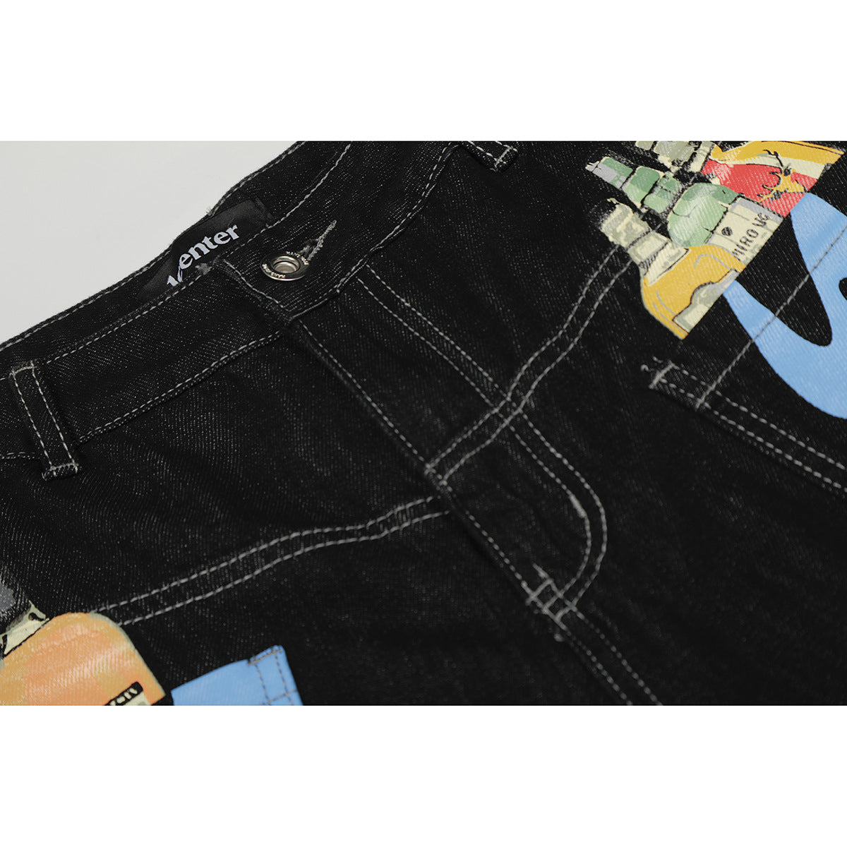 Close-up of black jeans showing the waistband, metal button, contrast stitching, and part of a colorful graphic.