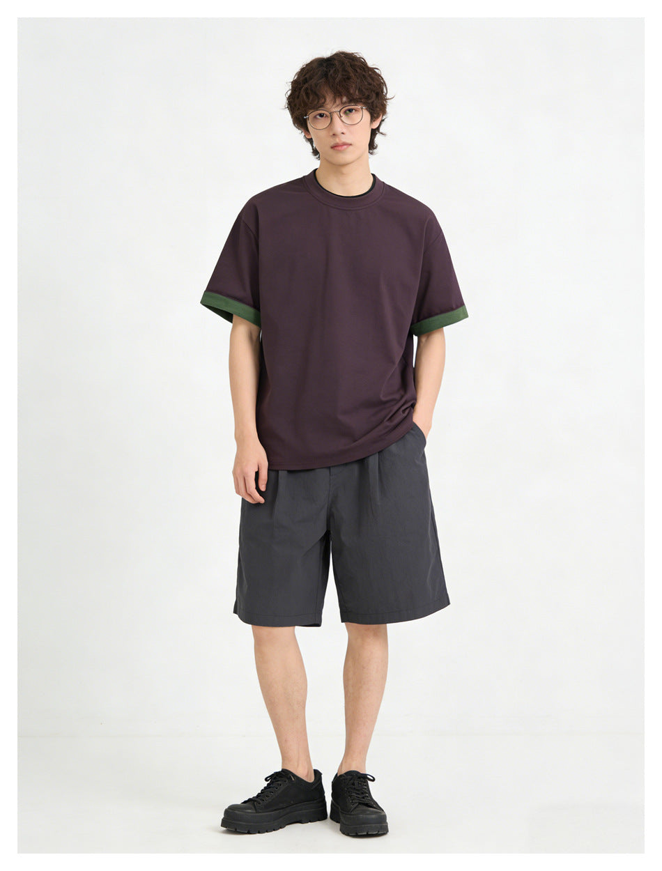 Front view of a model wearing a dark plum T-shirt with green sleeve cuffs and charcoal shorts.