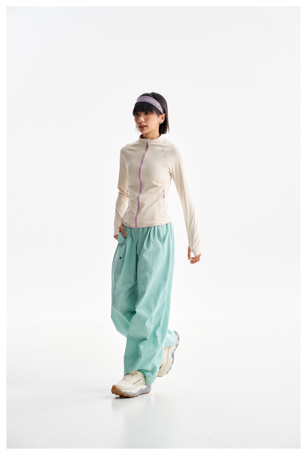 Model walking in an ivory zip jacket with lilac trim, loose aqua trousers, and white sneakers.