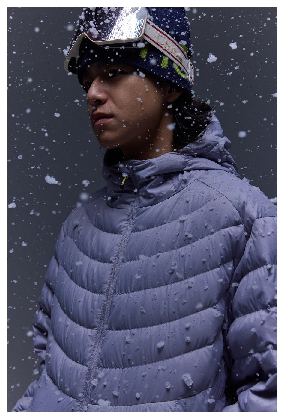 Close-up of a person wearing a lavender quilted puffer jacket and ski goggles as snow falls.