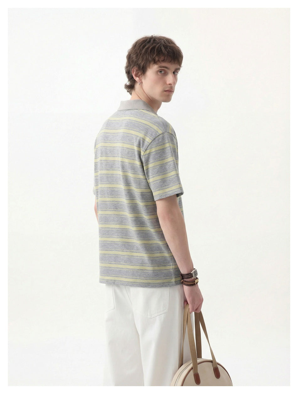 Back view of a man wearing a gray and yellow striped short-sleeve polo top with white pants, holding a tote bag